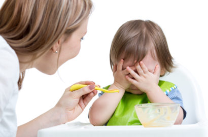 Mother trying to spoon feed her toddler who has his hands in front of his whole face as if he is refusing to eat