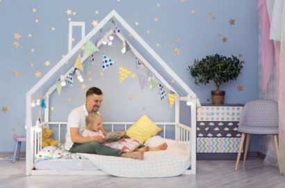 Father and 3 year old daughter in a framed toddler bed with the mattress at floor level. Stars on the wall and colourful flags attached to the frame of the bed which goes up and over the childs head in the shape of a house.